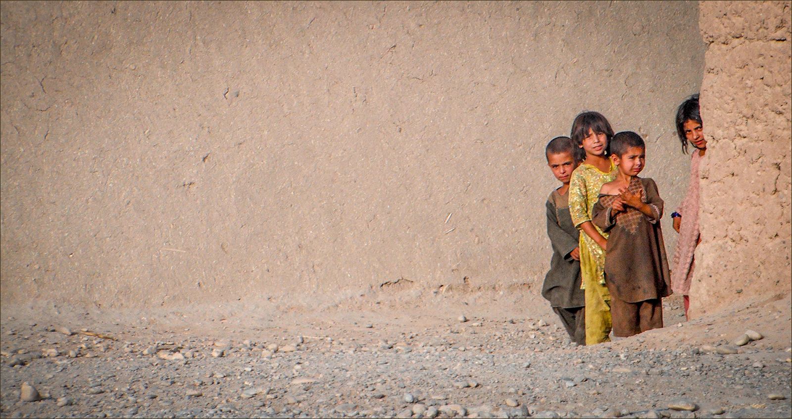 Four children peeking around the corner of a mud wall in a dusty rural village.