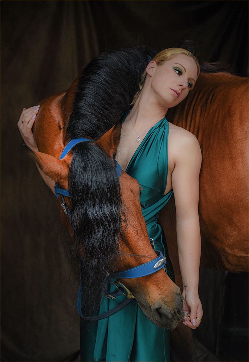 Woman in a teal dress leaning against a brown horse with its head resting on her shoulder.