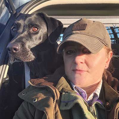Woman wearing outdoor clothing and a cap taking a selfie with a black dog inside a vehicle.