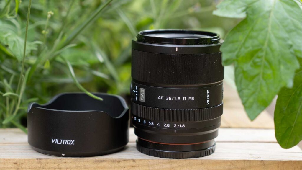 iltrox AF 35mm F1.8 II FE lens standing upright beside its petal-shaped lens hood on a wooden surface with green foliage in the background.