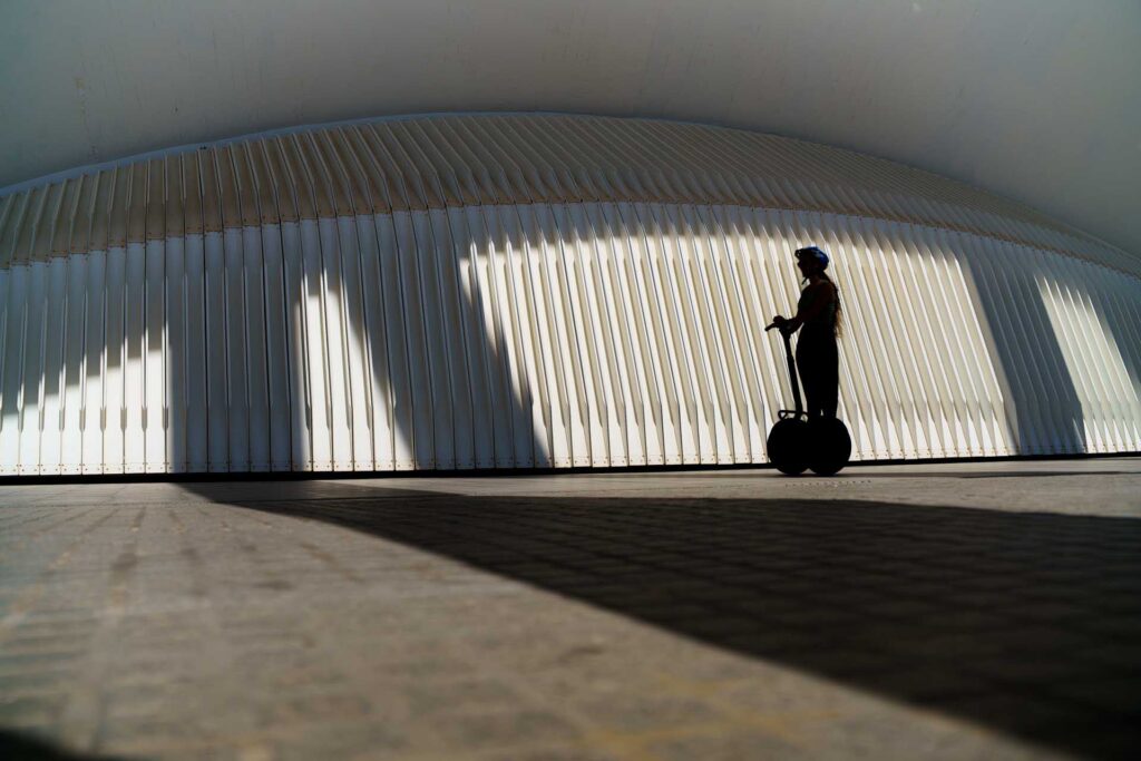 Silhouette of a person riding a Segway beneath Valencia's sweeping white modern architecture, with strong afternoon shadows stretching across the ground.