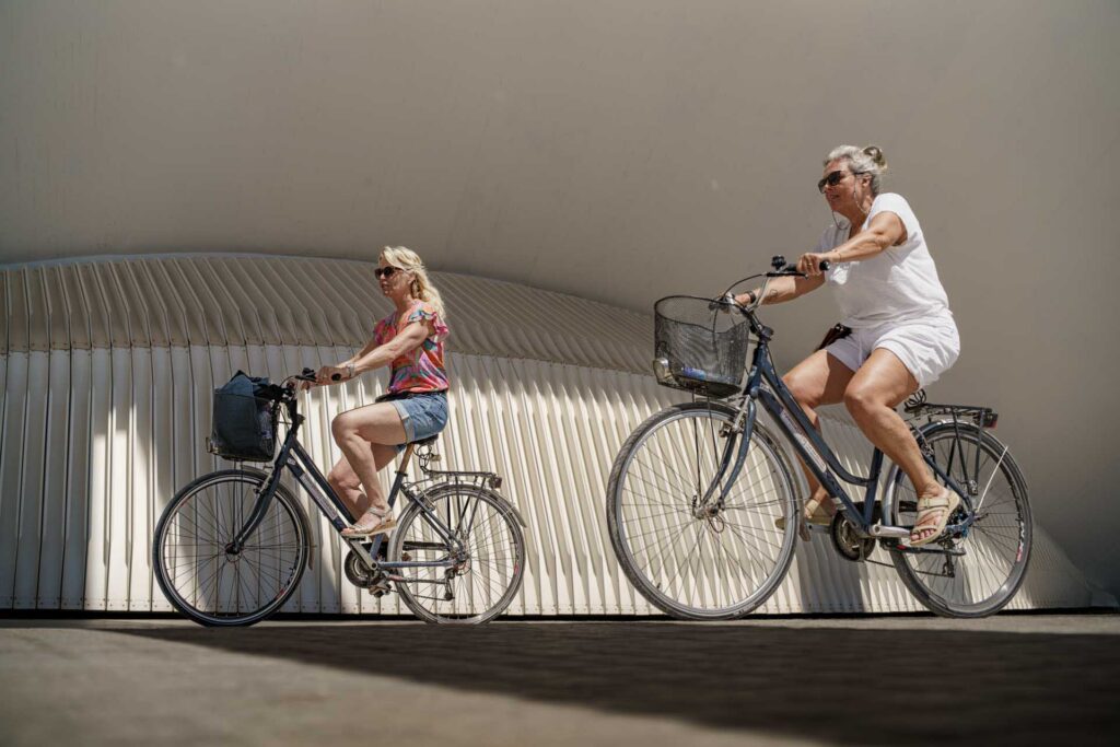 Two cyclists ride beneath Valencia's curved white architectural structure on a bright sunny day.