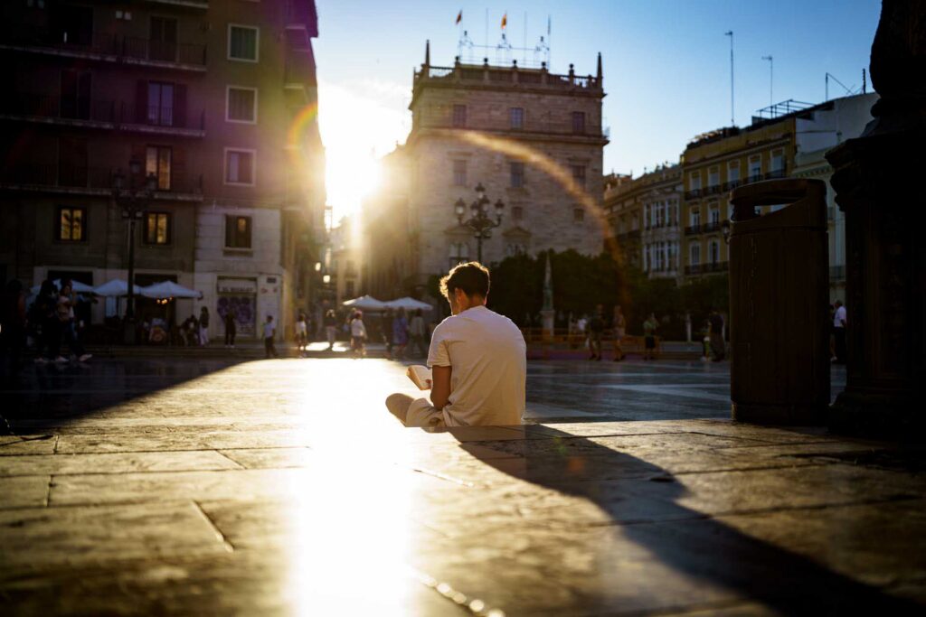 A person sits reading in a sunlit square in Valencia, with historic buildings, lens flare and long evening shadows behind them.