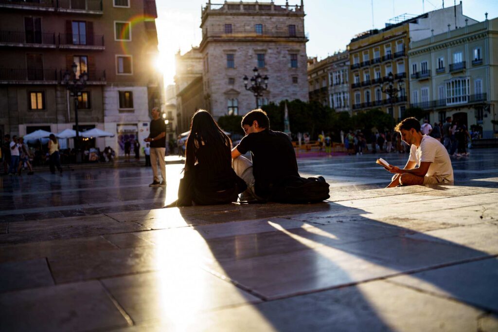 Three people sit chatting while another person reads alone in Valencia's Plaza de la Virgen at golden hour.