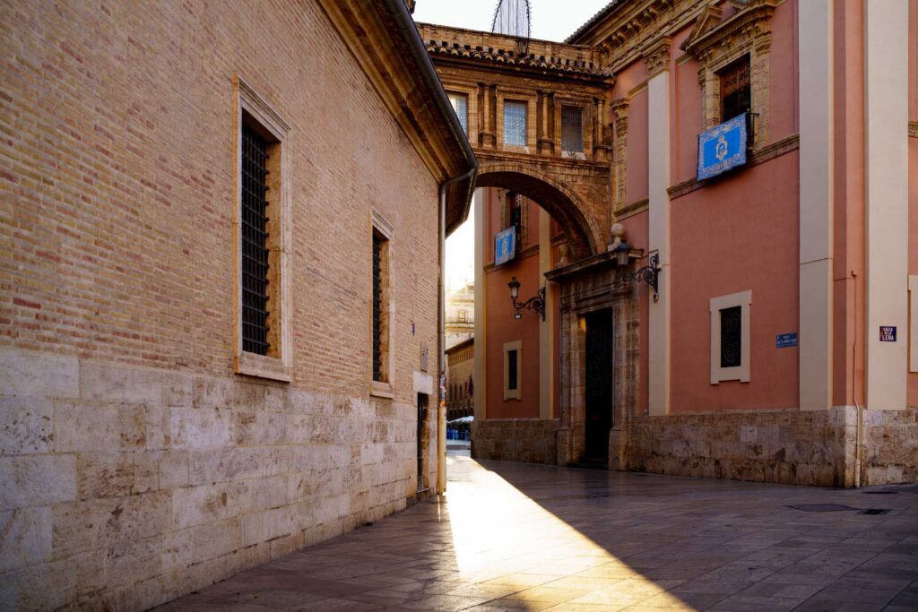 A quiet street in Valencia with historic buildings connected by an arched bridge, illuminated by low evening sunlight.