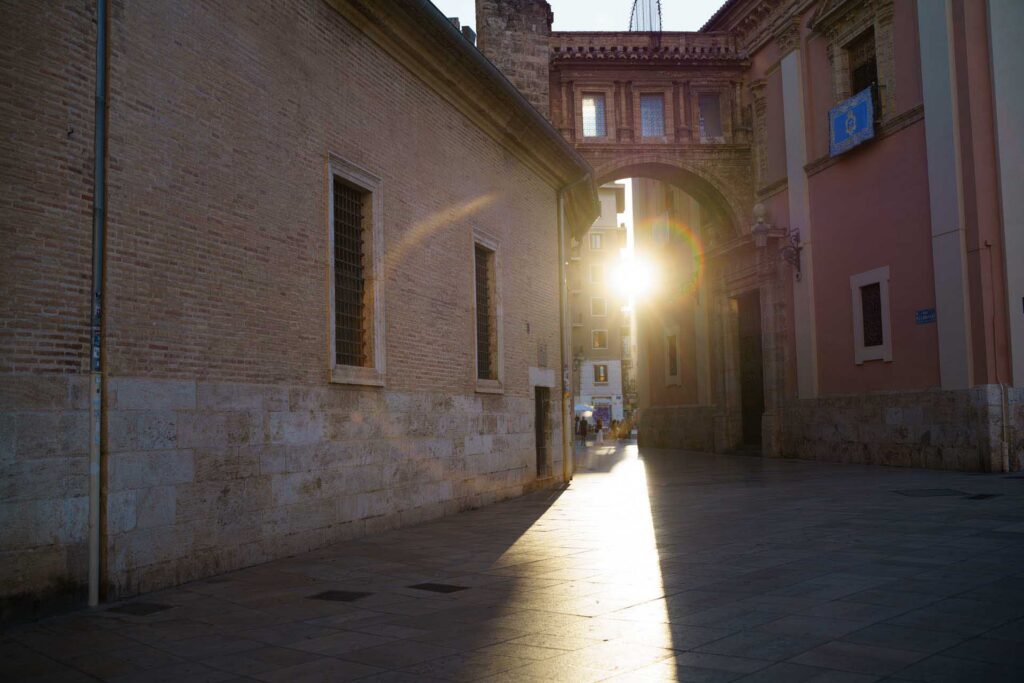 Sunlight shines through an arched passage between historic buildings in Valencia, creating a prominent lens flare.