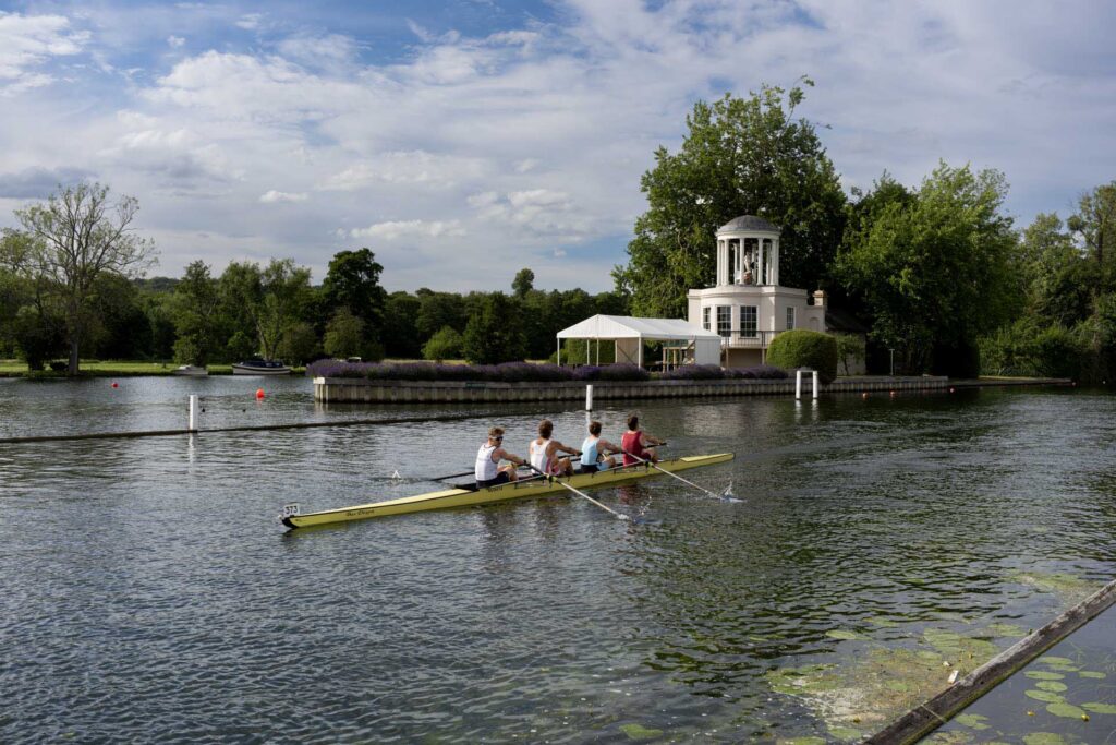 Four rowers paddle along the River Thames in Henley-on-Thames, with Temple Island and its white pavilion in the background.