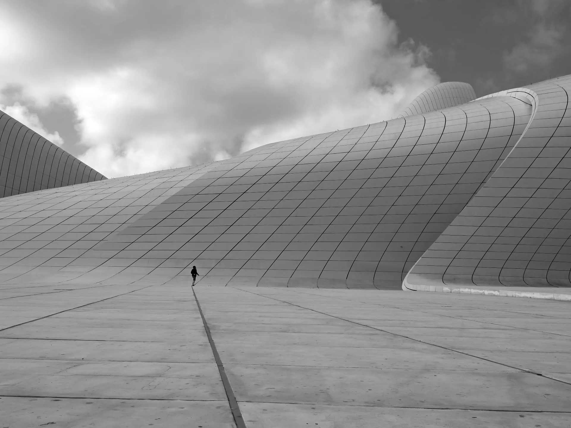 A lone person walks beside an immense curved building covered in geometric panels beneath dramatic clouds.