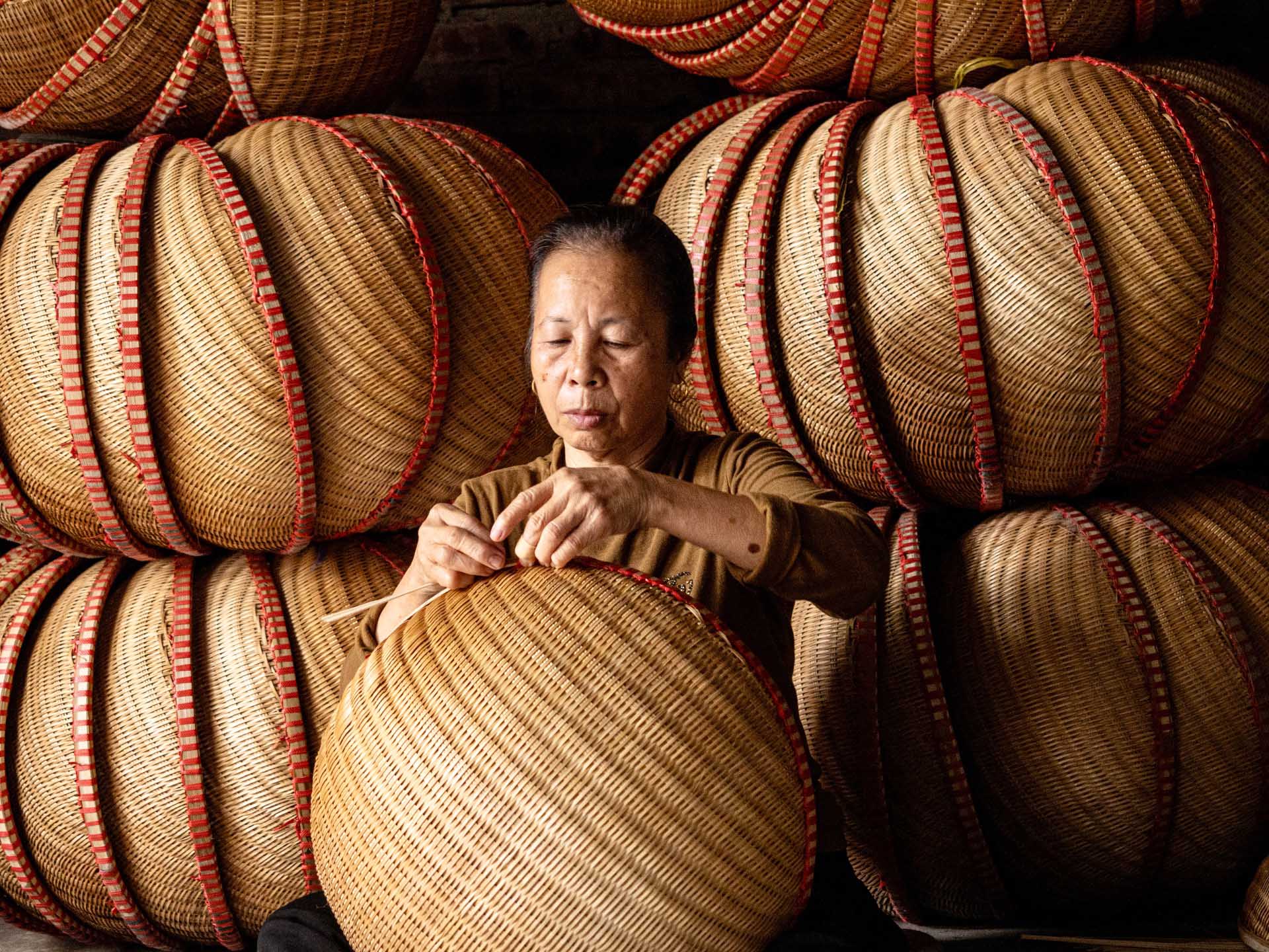 A woman works on a large woven basket while surrounded by stacks of rounded baskets edged with red binding.
