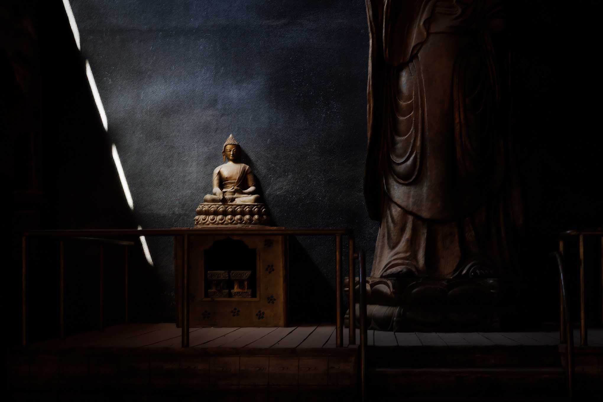 A small seated Buddha is illuminated by diagonal shafts of sunlight beside the feet of a much larger statue in a dark temple.