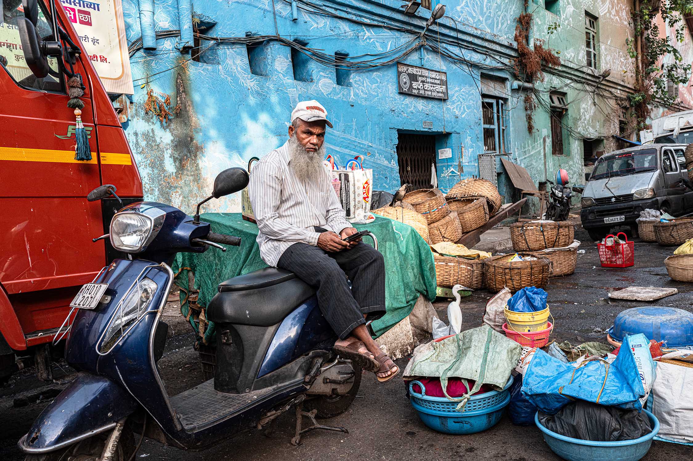 A bearded man sits on a blue scooter among baskets, bags and vehicles in a colourful market street with weathered blue walls.