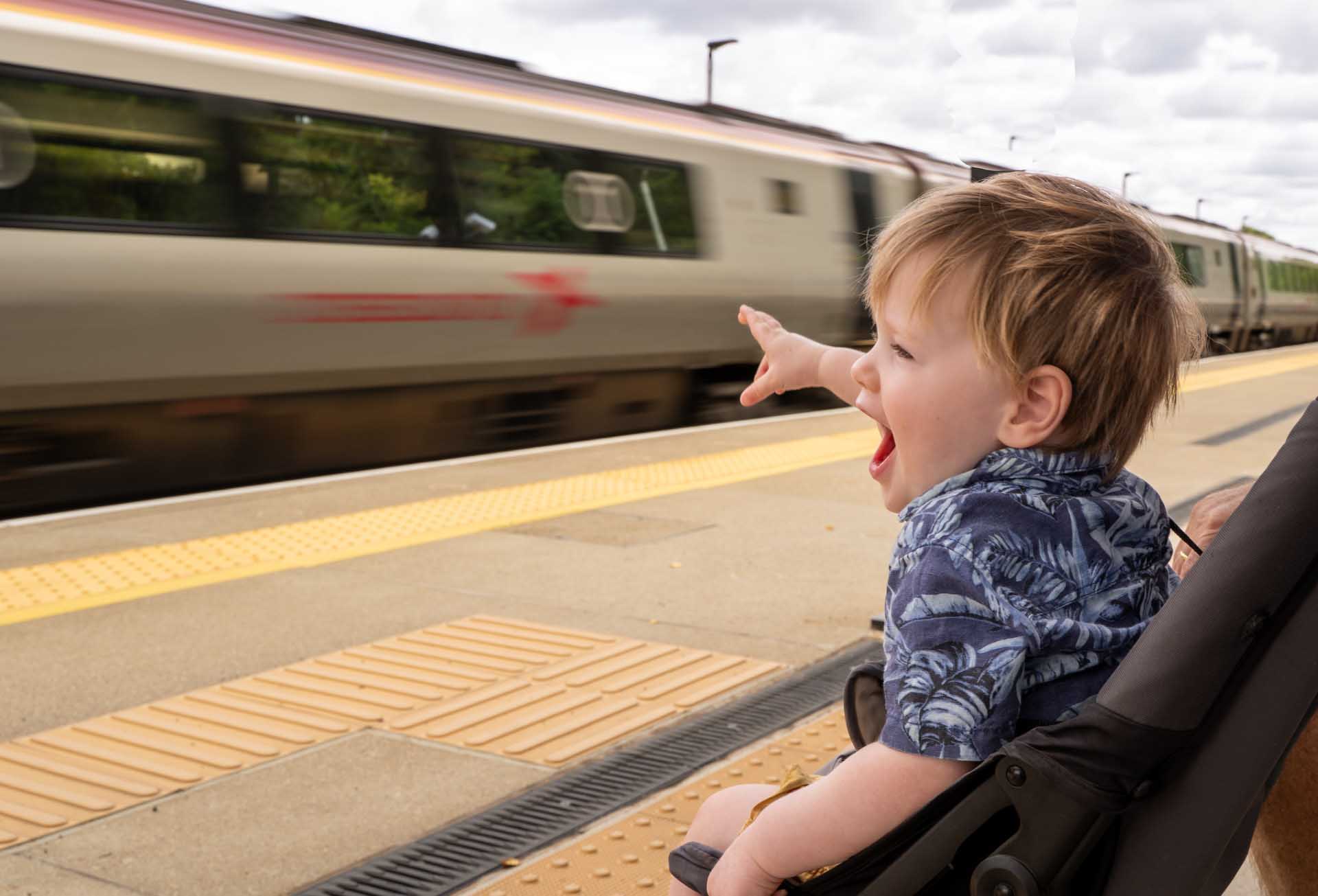 A toddler in a pushchair points excitedly at a train speeding past the station platform.