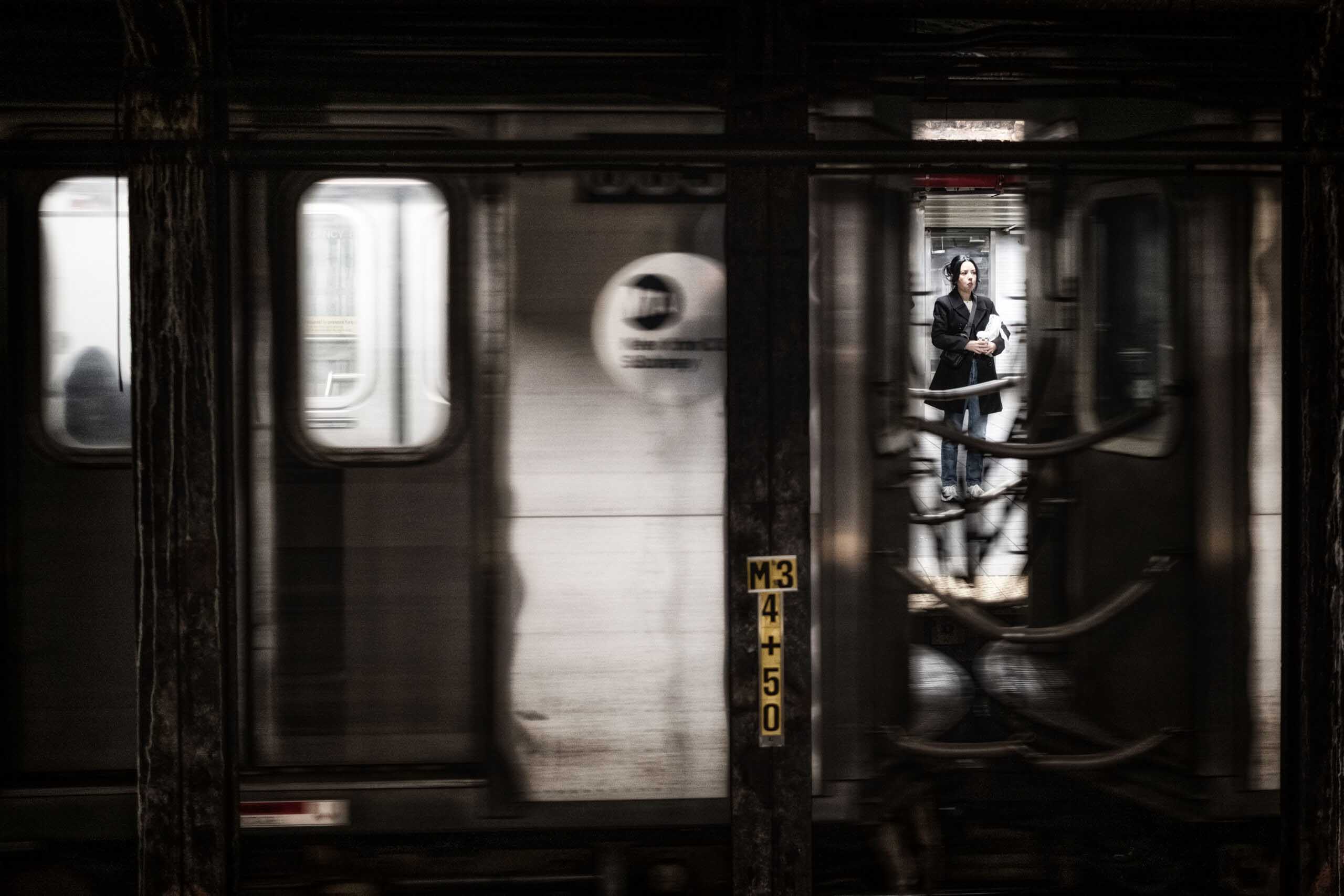 A woman waits on an underground platform, briefly framed through the windows of a motion-blurred subway train.