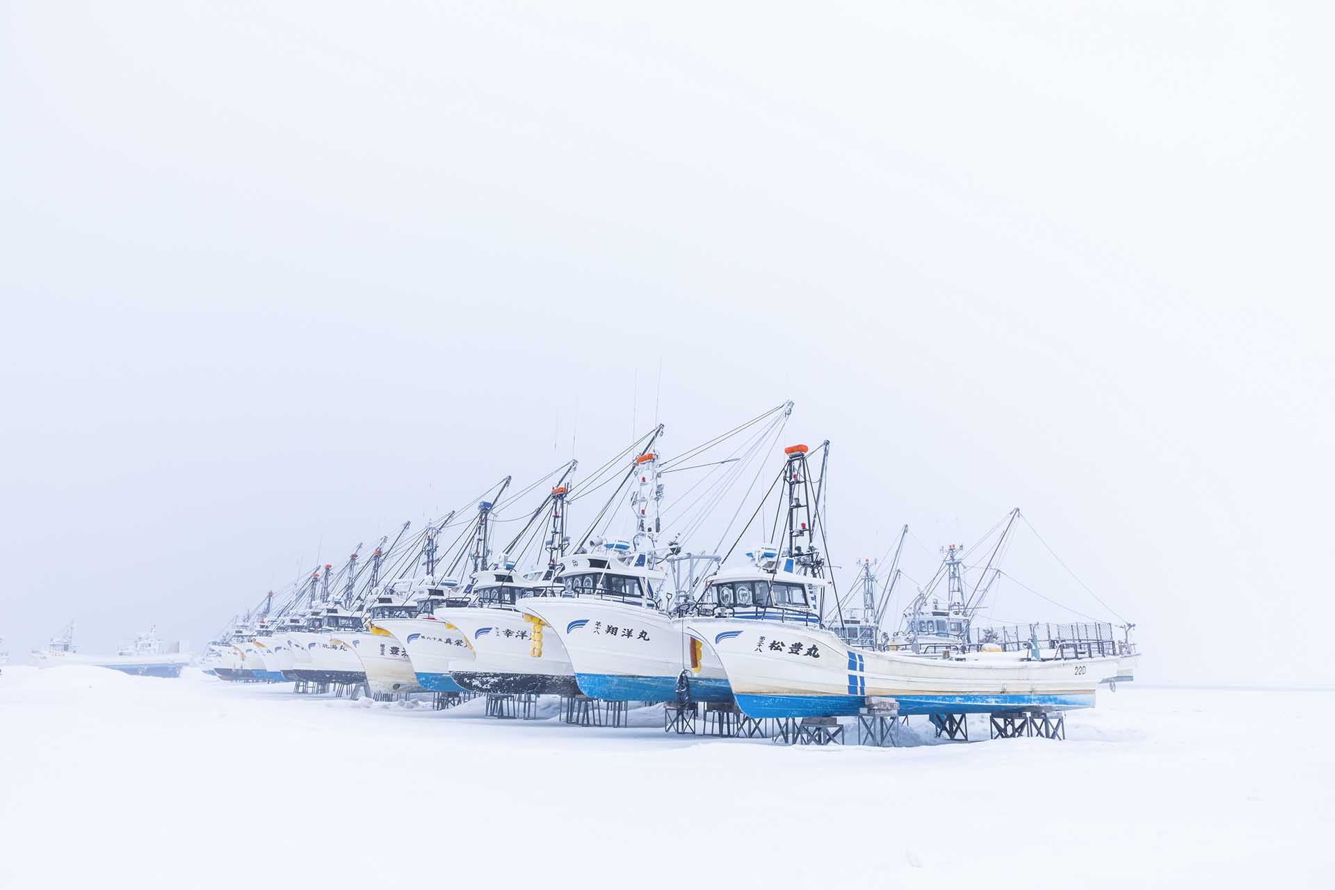 Rows of Japanese fishing boats rest on raised stands in deep snow, their masts disappearing into the pale winter sky.