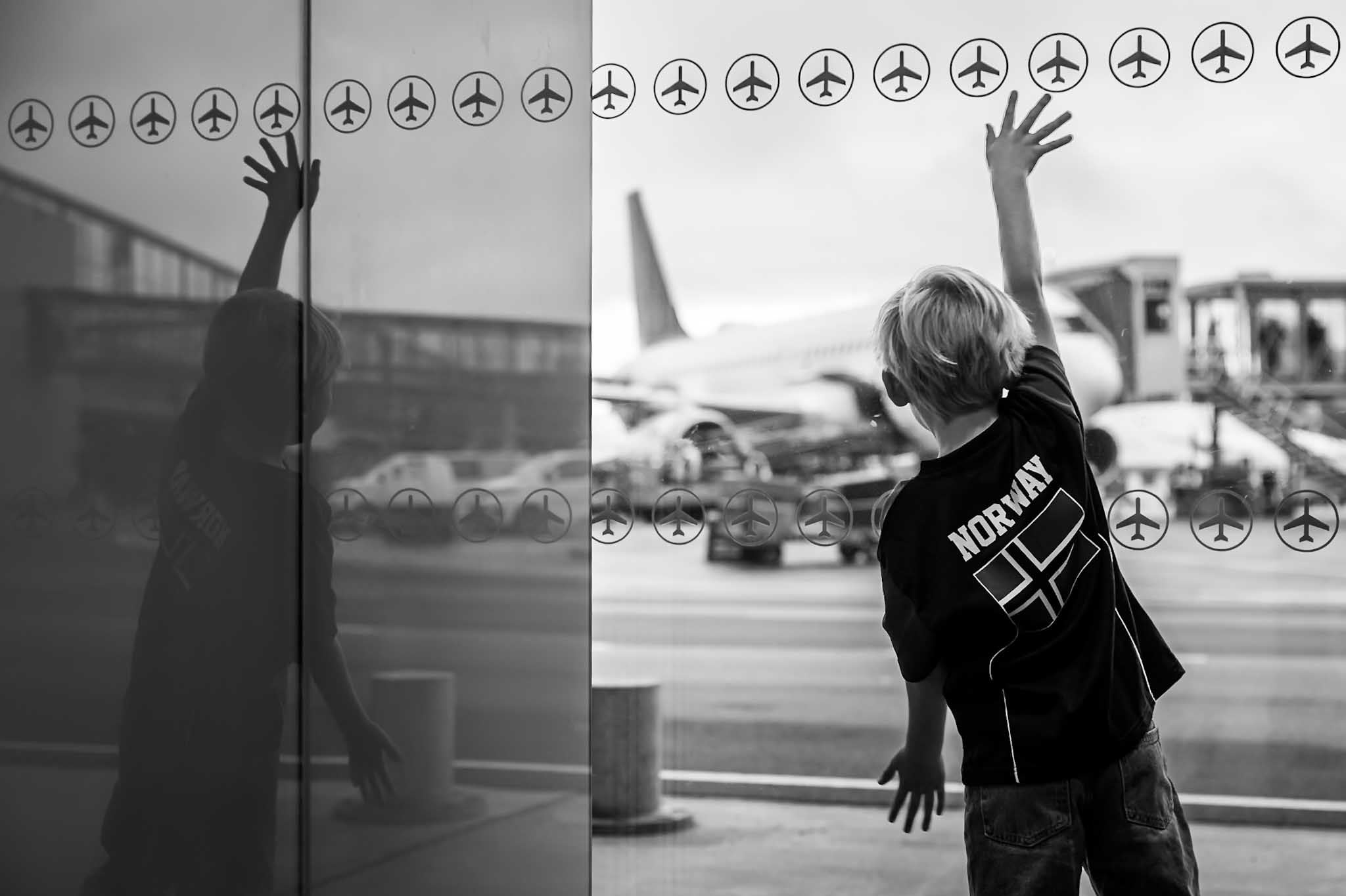 A child wearing a Norway shirt reaches towards aeroplane symbols on an airport window, with a passenger aircraft outside.