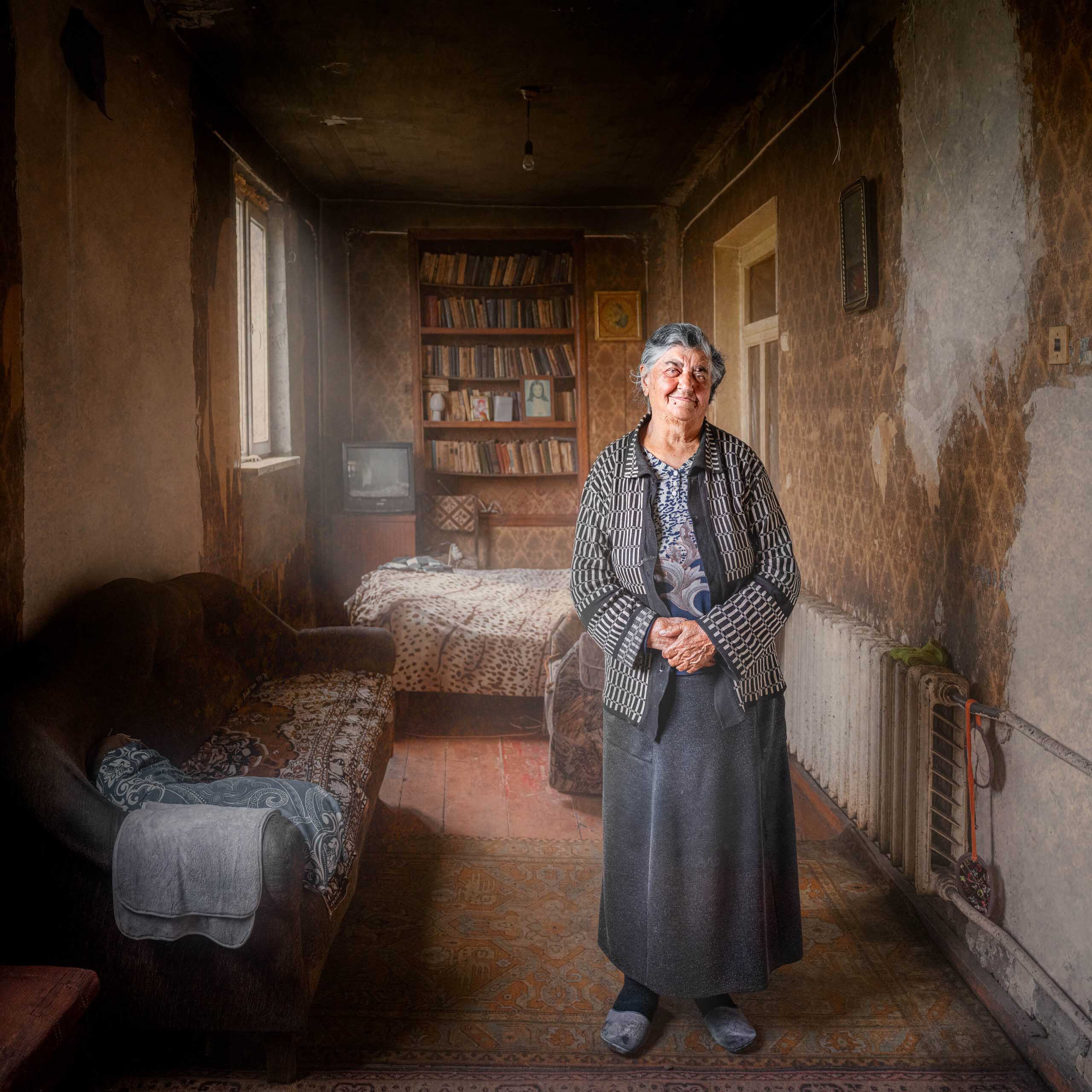 An elderly woman smiles in a timeworn room furnished with a sofa, bed, bookcase, television and radiator.
