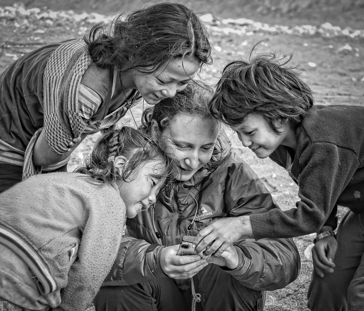 Four people gather closely around a compact camera, smiling as they review a photograph in a black-and-white outdoor scene.