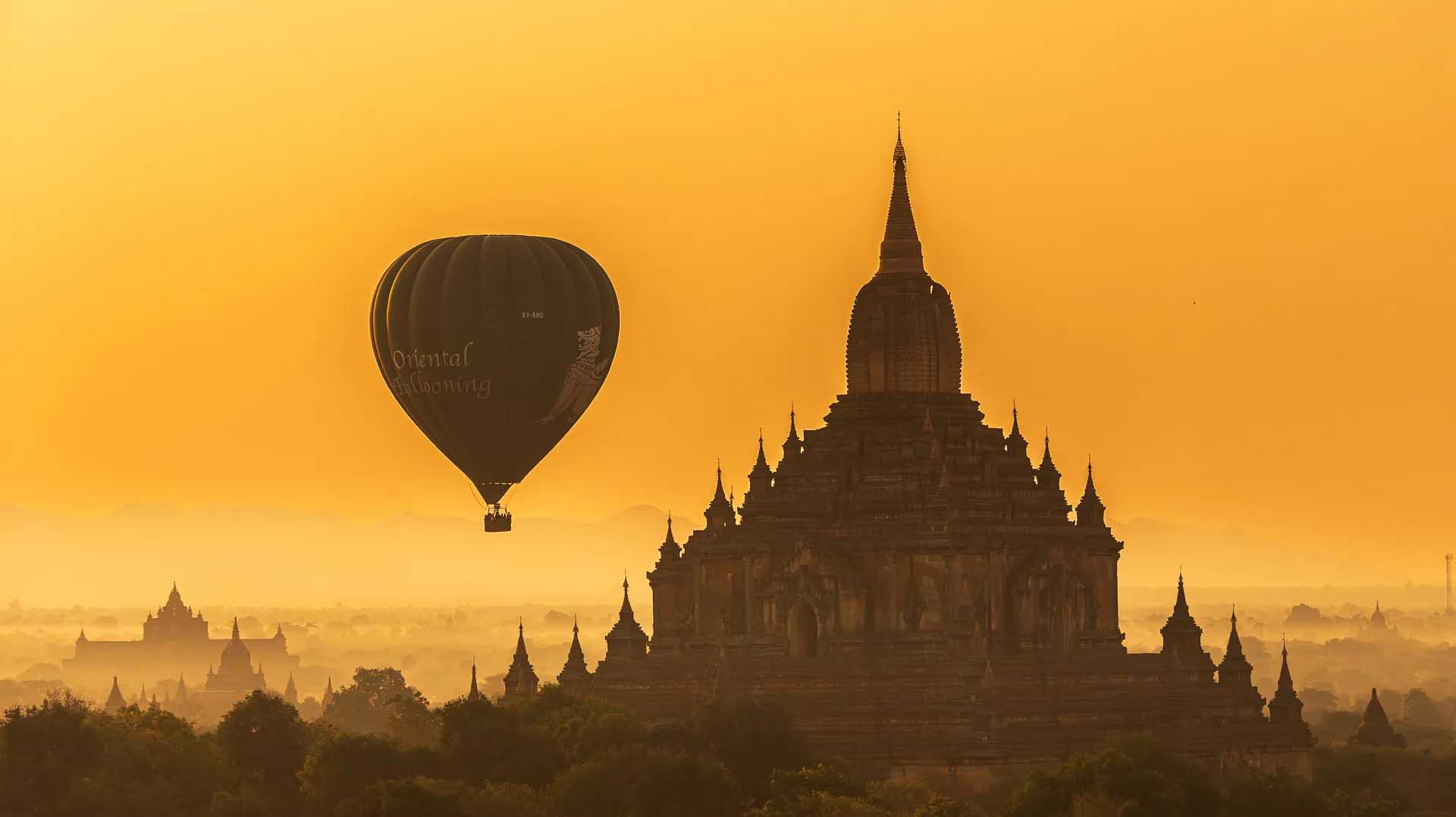 A hot-air balloon marked “Oriental Ballooning” floats beside the silhouette of a temple above Bagan’s misty landscape at sunrise.
