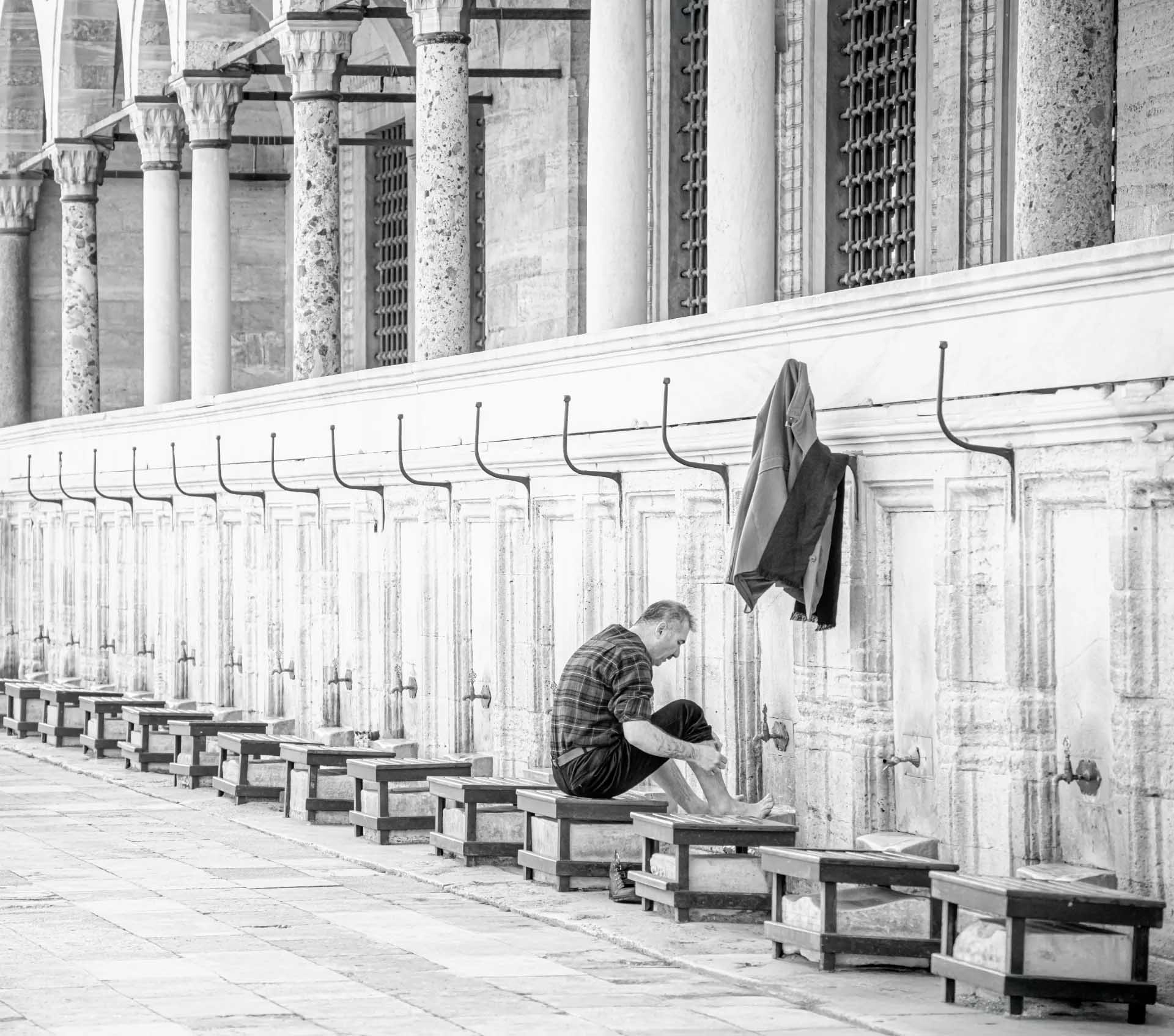 A man washes his feet at a long row of taps beneath stone columns, with his jacket hanging from a hook above.