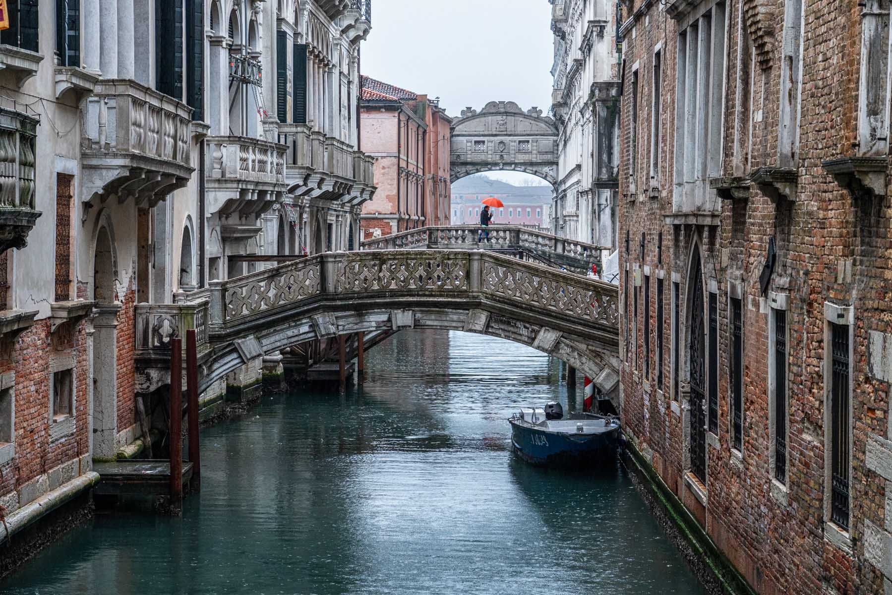 Rain falls on a narrow Venetian canal crossed by two stone bridges, while a person carrying a red umbrella walks in the distance.