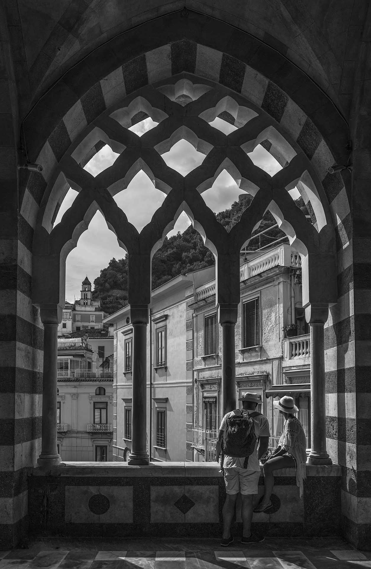 Two visitors look across a town from beneath an ornate striped arch with pointed openings and slender columns.