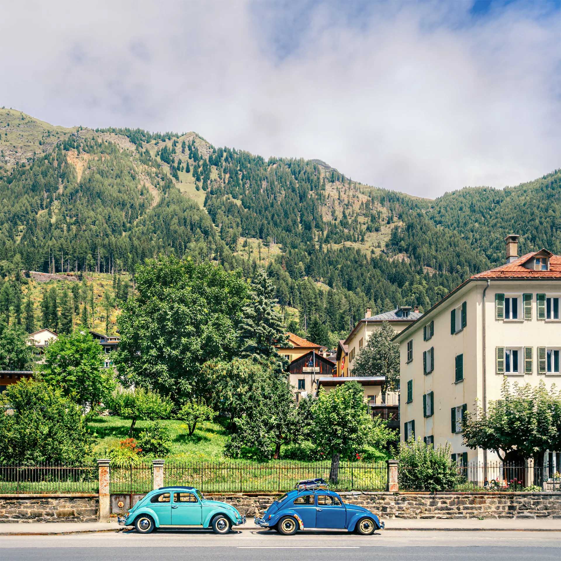 Turquoise and blue vintage Volkswagen Beetles parked beside a green garden, with a forested Alpine mountainside behind.