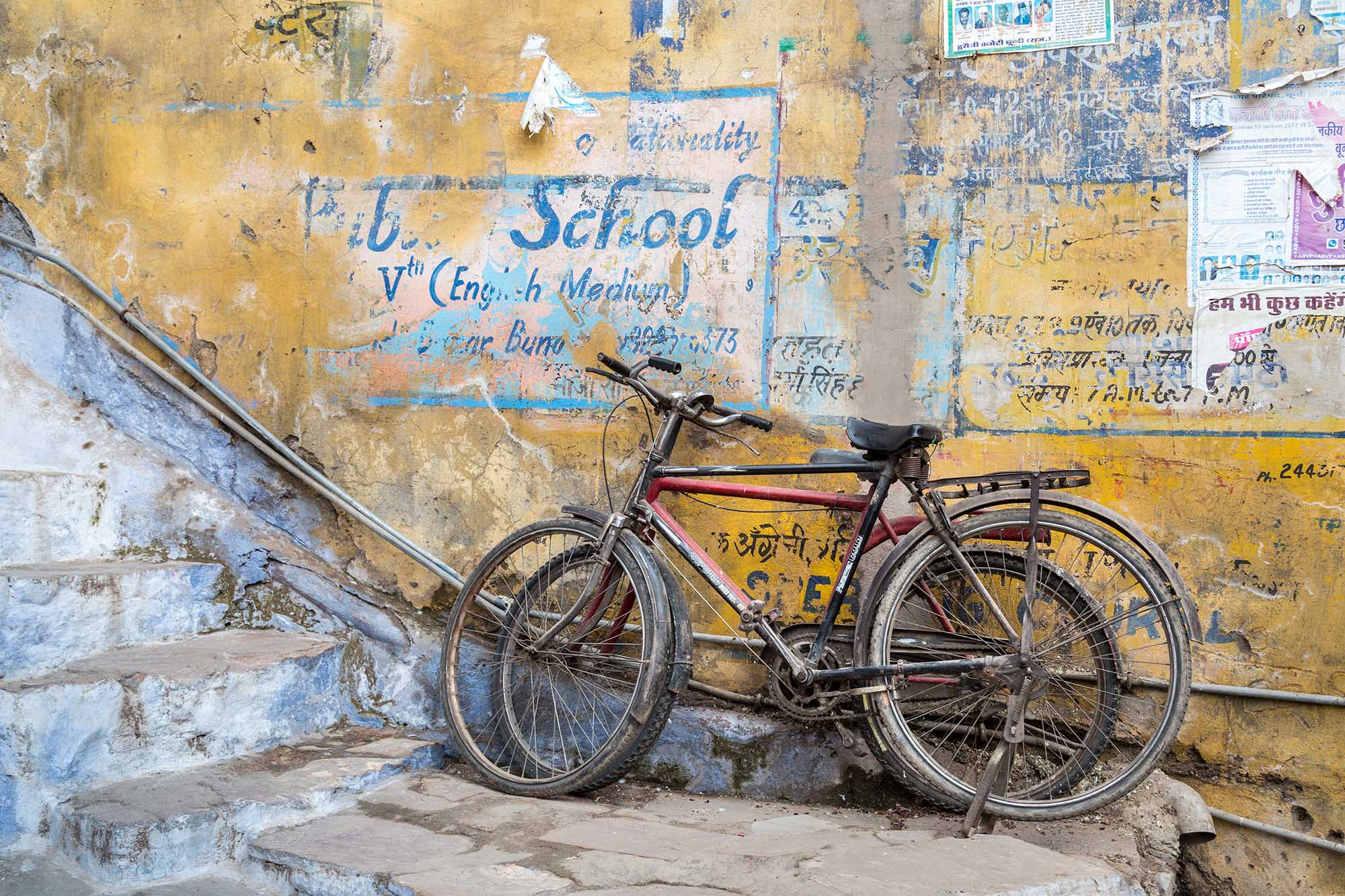 Two worn bicycles lean against a weathered yellow wall covered with faded school signs, handwritten notices and posters in Hindi and English.