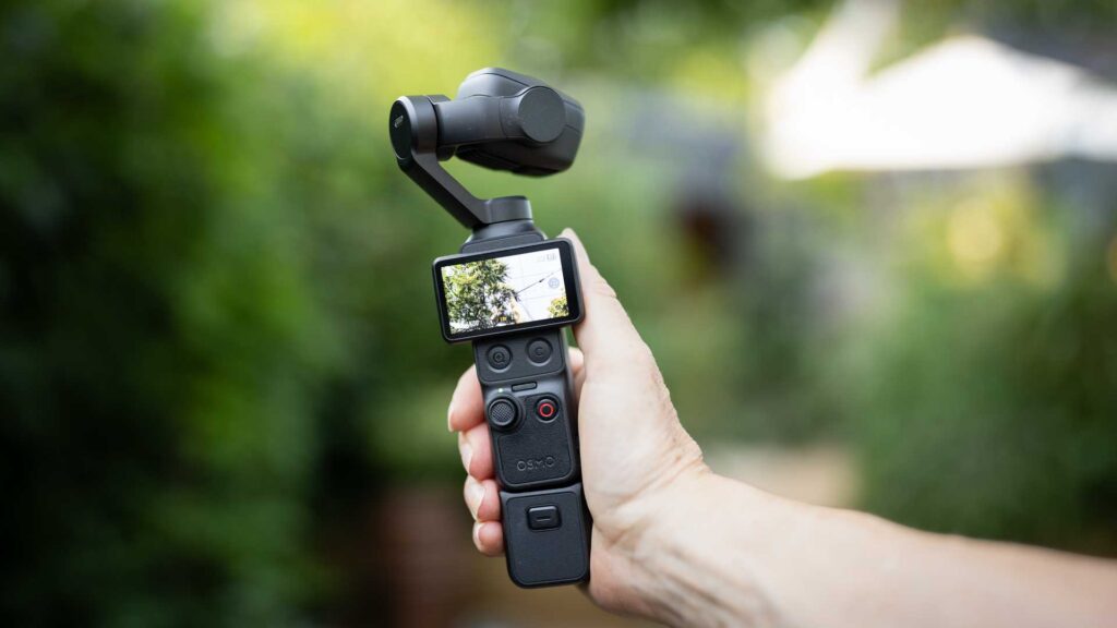 DJI Osmo Pocket 4P held upright in a garden, with the touchscreen, joystick and red-ringed record button visible.