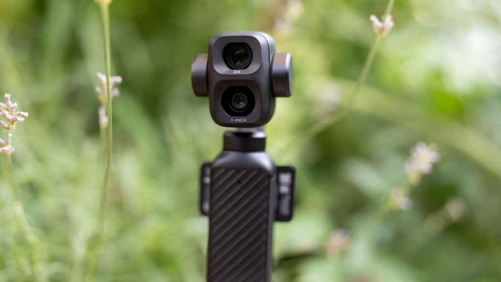 Close-up of the DJI Osmo Pocket 4P camera head, with lenses labelled “3X” and “1-INCH” surrounded by softly blurred garden plants.