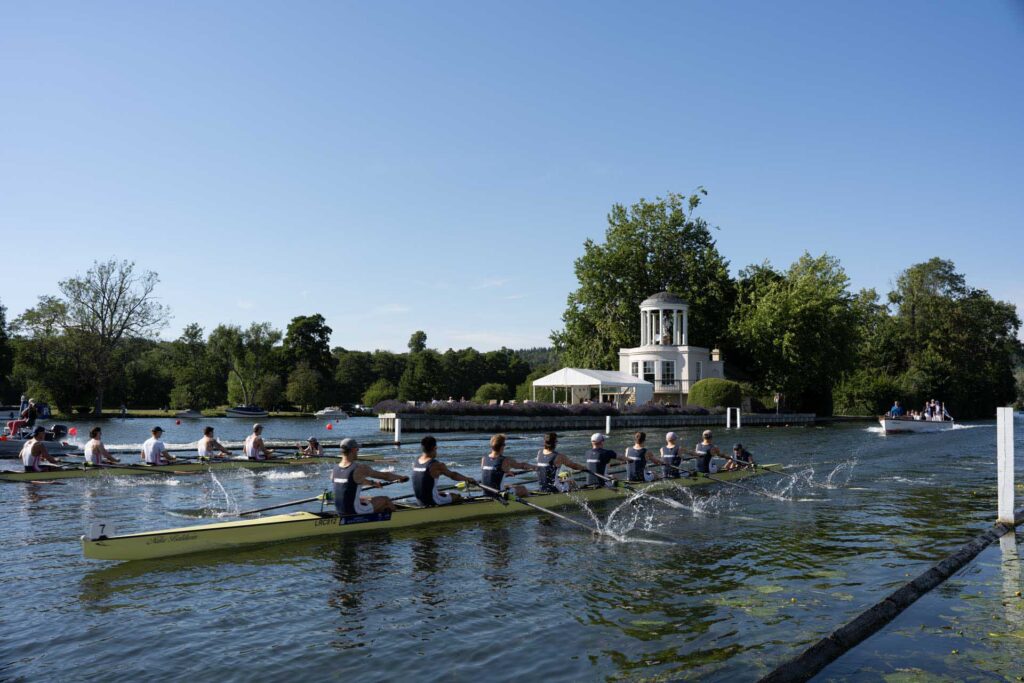 Two rowing crews race across a river near a white riverside pavilion, with a motorboat and tree-lined banks under a clear blue sky.
