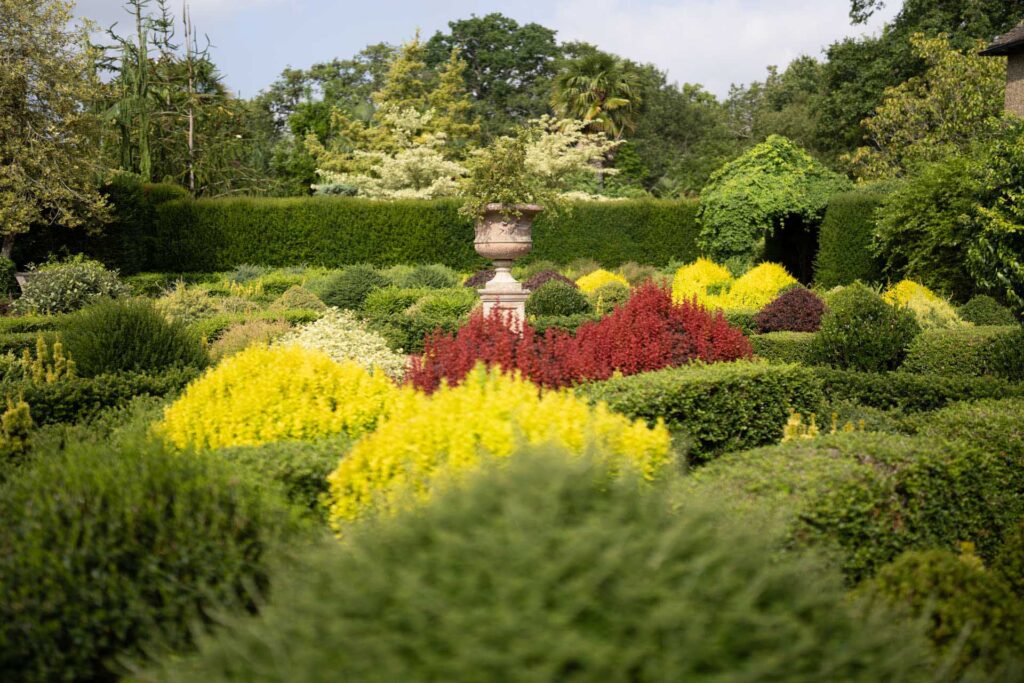 Stone urn centred among red, yellow and green planting in a formal garden, with a softly blurred foreground.