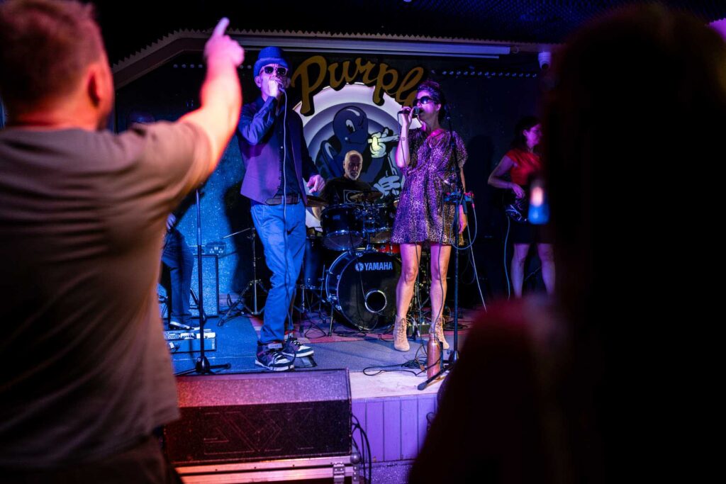 Band performing under blue and magenta lights while an audience member raises an arm in the foreground.