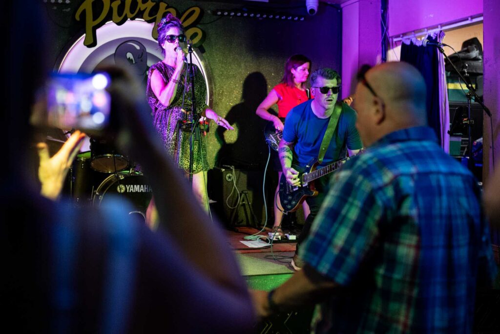 Singer and guitarist performing under purple and green lights as audience members watch and record on a phone.