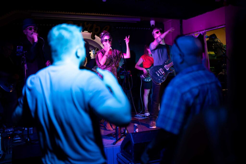 Singers, guitarist and bassist performing beneath blue and magenta lighting, framed by two audience members.