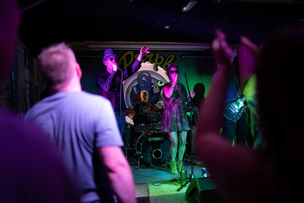 Two singers perform in front of a drummer under purple and green stage lights as raised hands frame the scene.