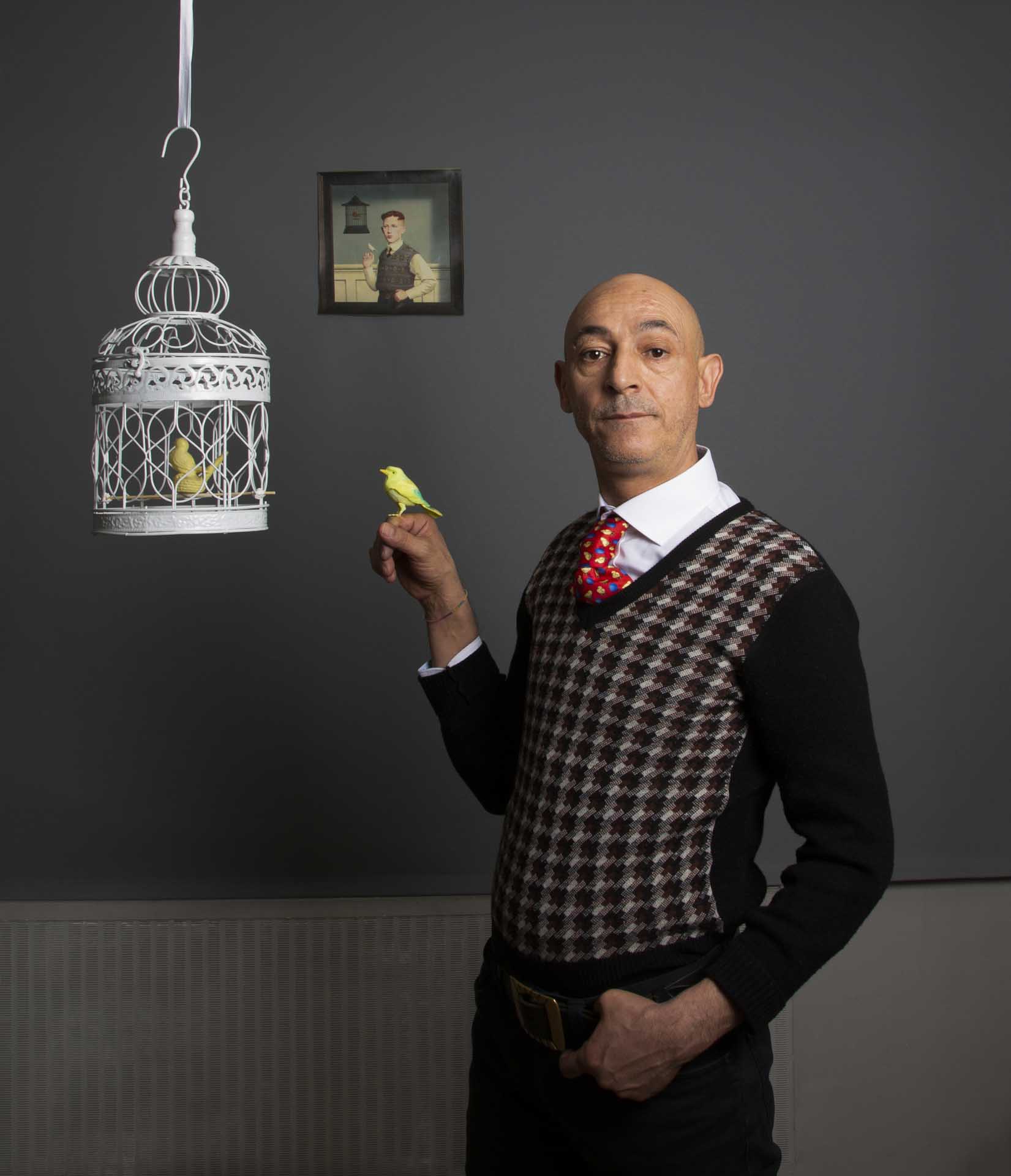 Bald man in a patterned sweater vest and bright red tie holds a yellow bird beside an ornate white birdcage, set against a dark grey wall with a small framed portrait.