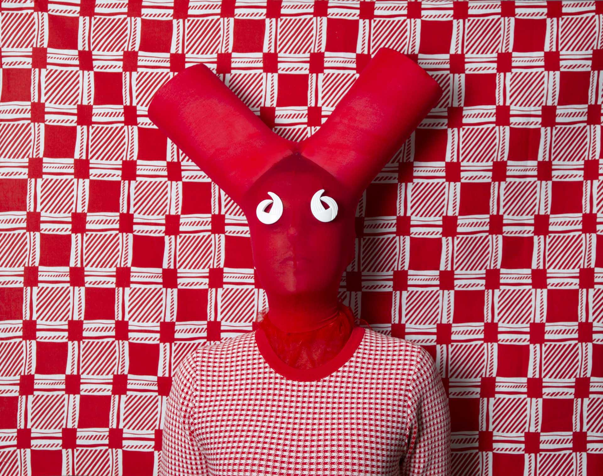 Person wearing a sculptural red Y shaped mask with white curled eyes stands against a bold red and white checked backdrop.