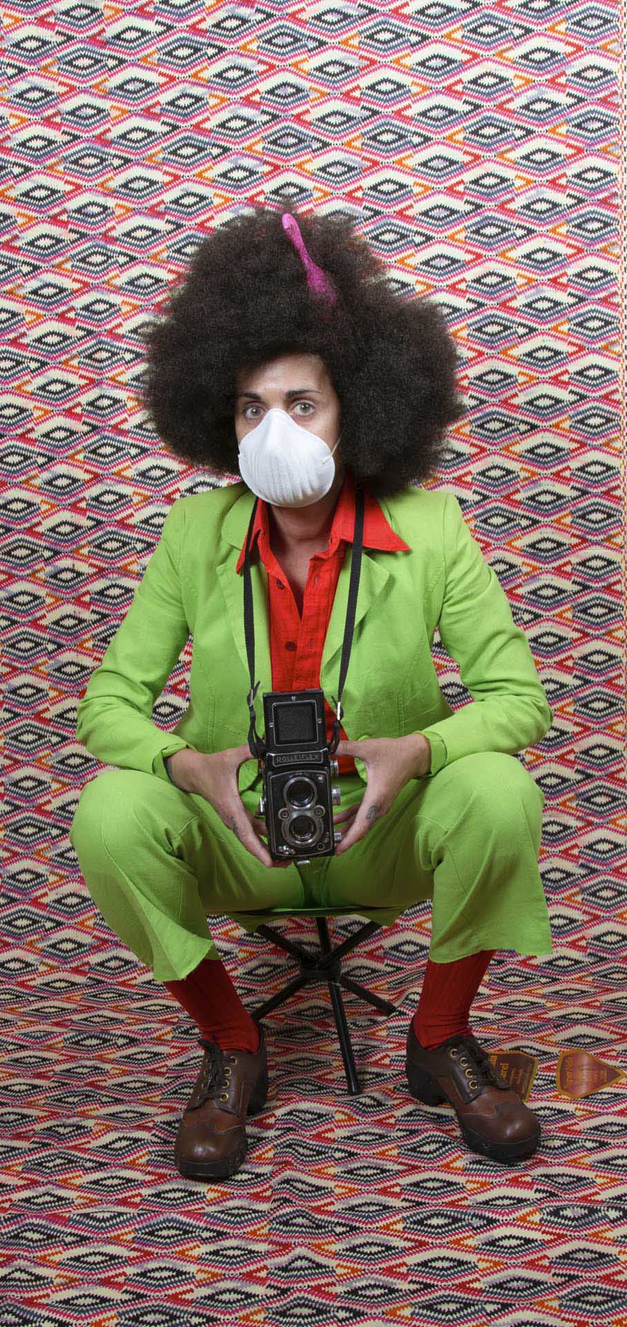 Person with a large afro wears a white respirator mask, lime green suit and red shirt while holding a twin lens camera against a multicoloured geometric backdrop.