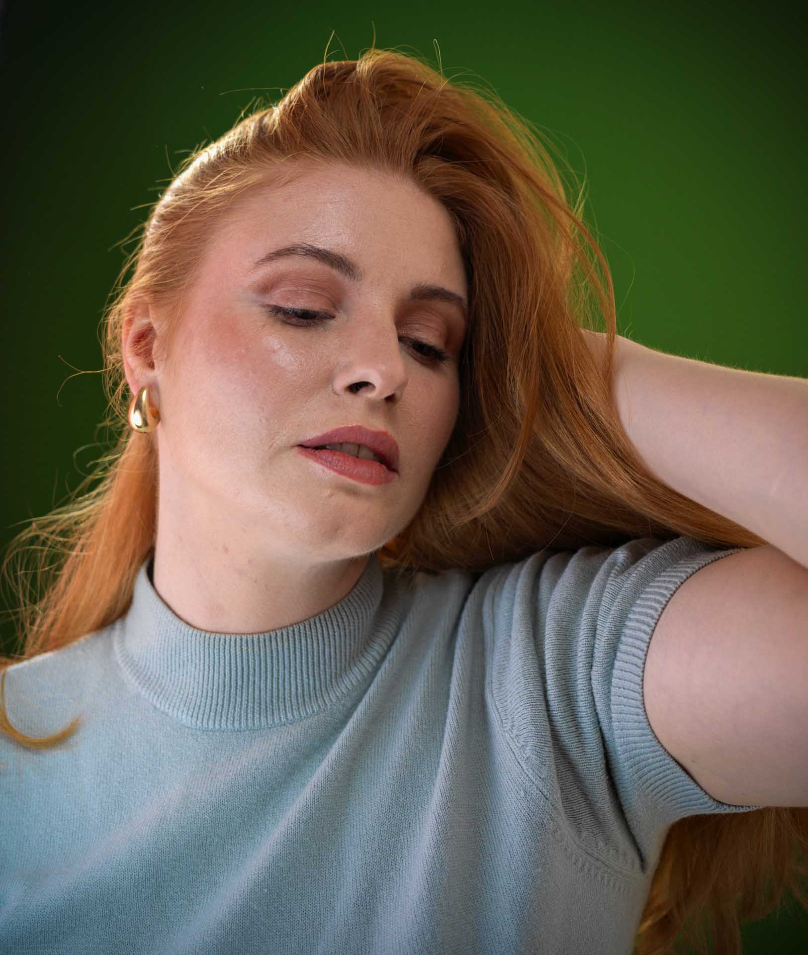 Close portrait of a red haired woman in a pale blue knitted top, resting one hand behind her head against a deep green background.