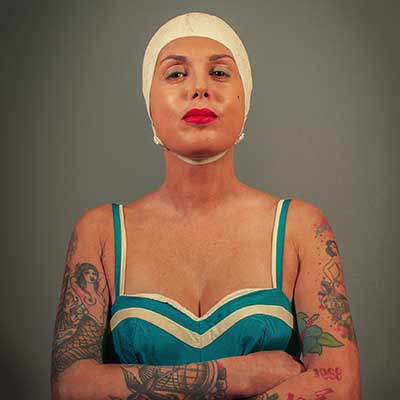 Chris Pig poses with folded arms against a grey background, wearing a white cap, red lipstick and a teal dress with cream trim, with colourful tattoos covering both arms.