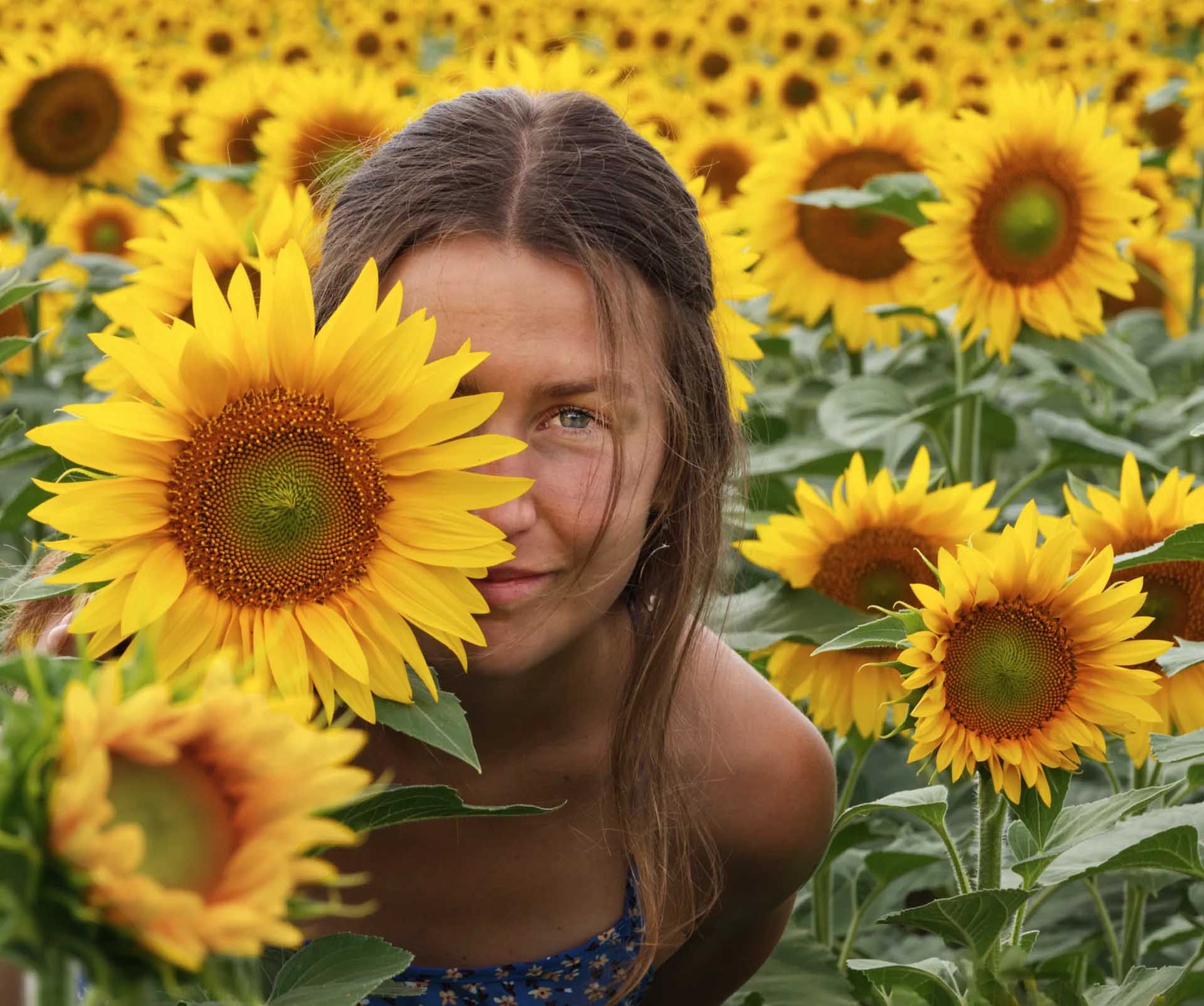 Young woman looking towards the camera from among yellow sunflowers, with one large bloom partly covering her face.