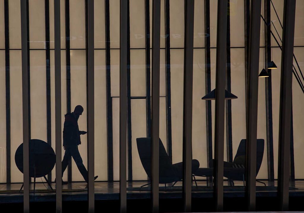 A silhouetted person walks past lounge chairs behind tall vertical window frames in a softly lit modern interior.