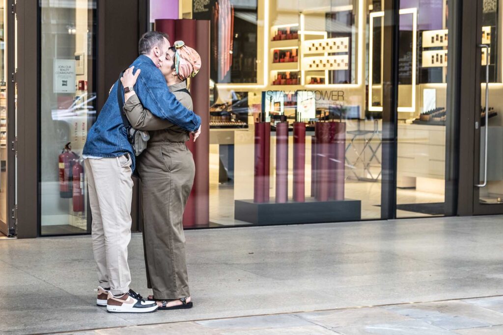 Two people embrace outside a brightly lit cosmetics shop, one wearing a patterned headscarf.