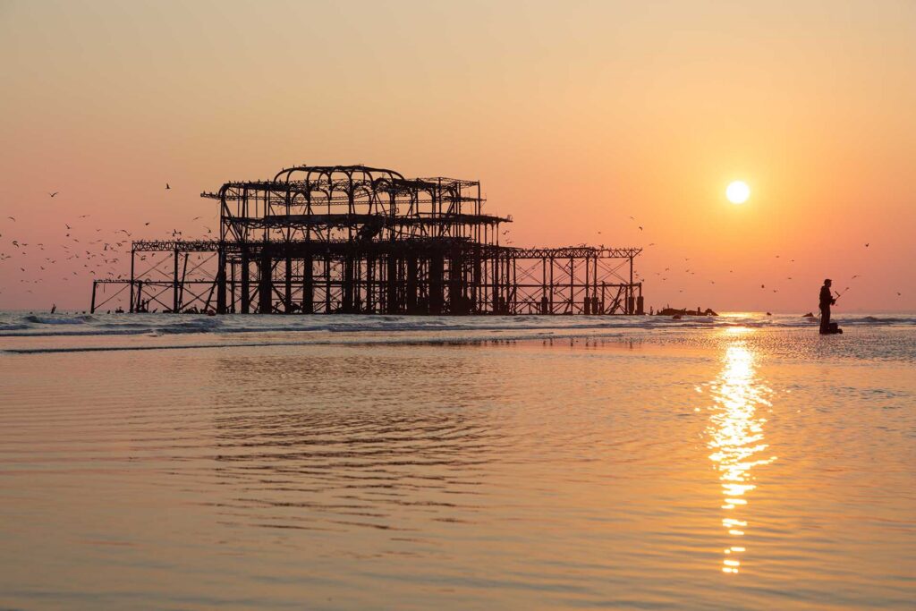 Brighton's ruined pier silhouetted against an orange sunset, with seabirds overhead and a lone angler standing in shallow water.