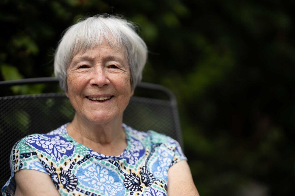 Smiling older woman with short white hair seated outdoors against a dark, softly blurred green background.