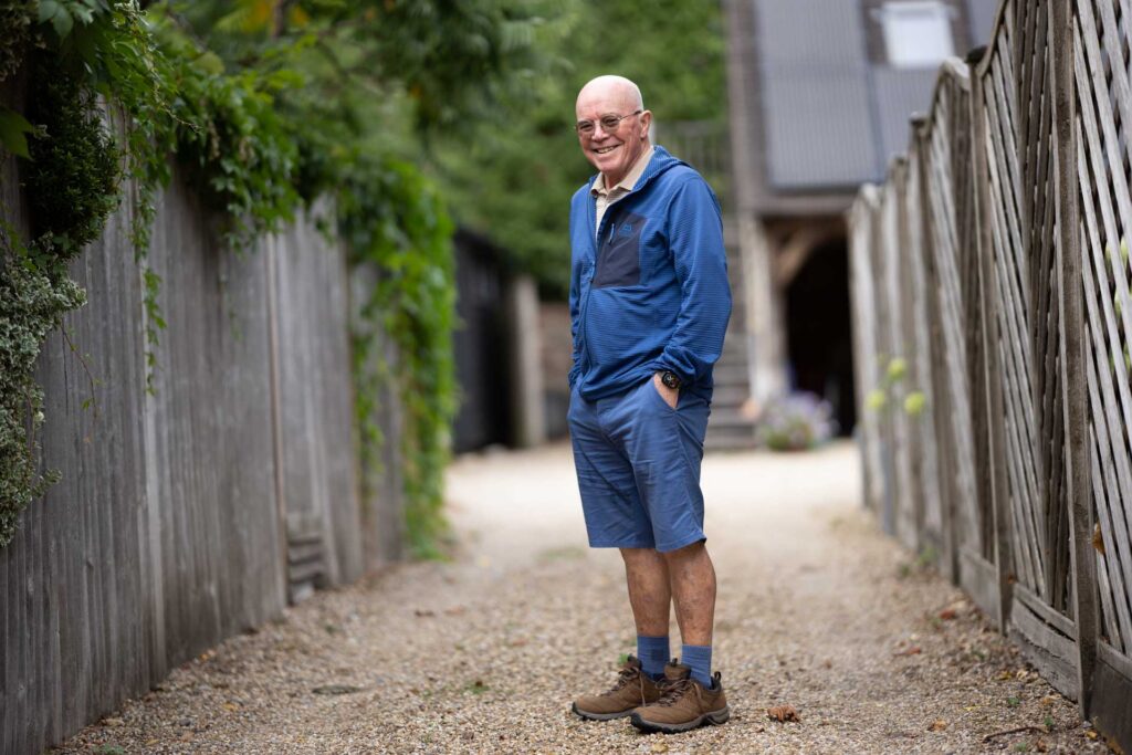 Smiling older man in a blue jacket and shorts standing on a gravel path between wooden fences.