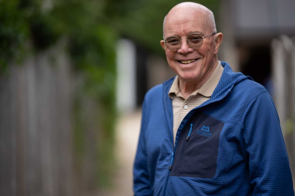Close portrait of a smiling older man wearing round glasses and a blue jacket, with a strongly blurred outdoor background.