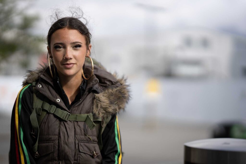 Young woman wearing large hoop earrings and a fur-trimmed vest, photographed outdoors against a softly blurred background.