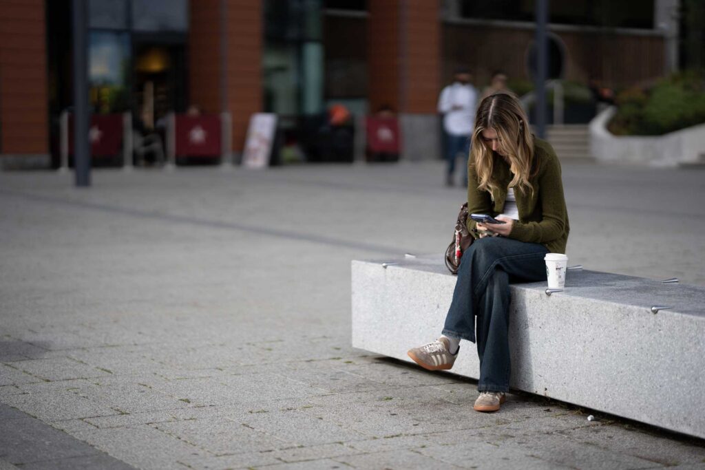 Young woman sitting alone on a concrete bench looking at her phone, with a takeaway cup beside her in an open plaza.