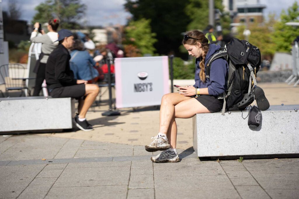 Young woman with a large hiking backpack sitting on a concrete bench and looking at her phone in bright sunshine.